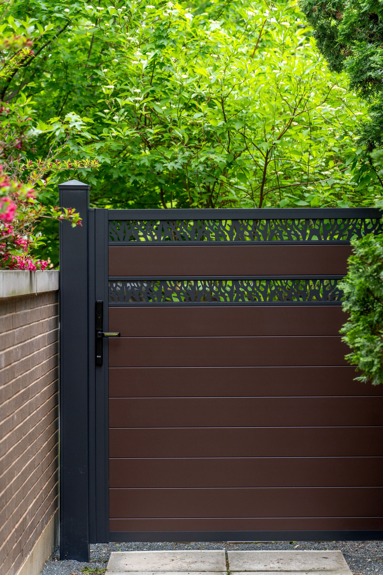 A premium real-world installation of a modern modular single-swing aluminum walk gate featuring a heavy-duty matte black frame and post with an integrated smart handle lock. The gate showcases a monotone deep architectural brown aluminum privacy board infill system accented by two stacked decorative organic lattice panels, beautifully integrated alongside a sleek brick wall and lush green landscaping.