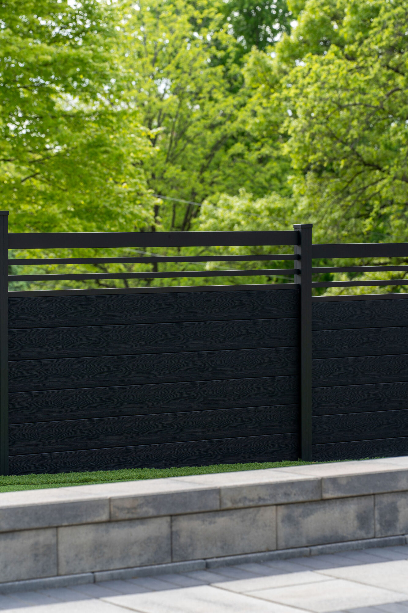 GWF® Classic Trellis charcoal composite fence panel straight on above a stone paver wall, open trellis crown of slim matte black rails over woodgrain infill