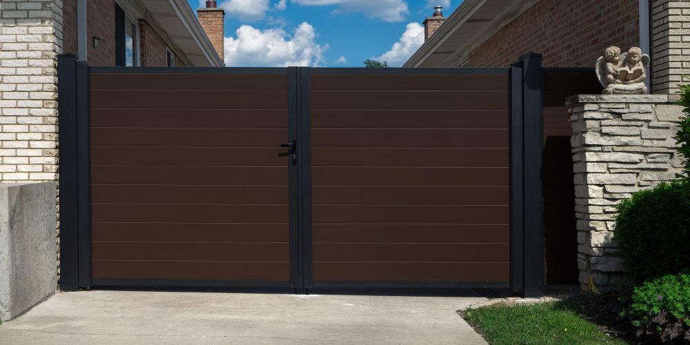 A cropped, high-end architectural installation of a modern modular double-swing aluminum driveway gate featuring a matte black frame and a matching central latch mechanism. The gate utilizes premium interlocking aluminum privacy board infills in a deep walnut brown finish, spanning a wide concrete driveway alongside a light stone masonry building wall.